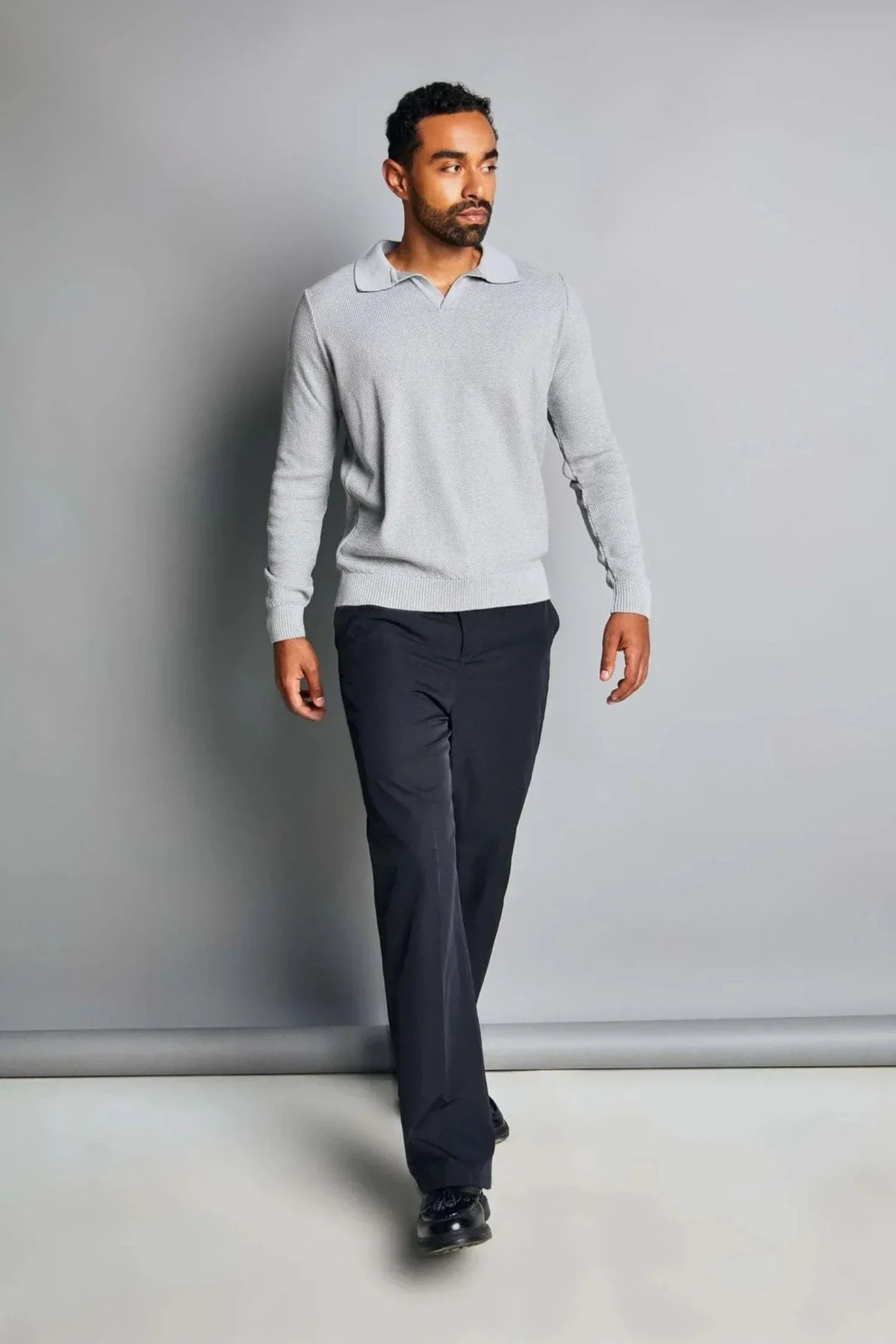 Hombre con suéter gris de punto y pantalón oscuro en fondo neutro, moda masculina elegante